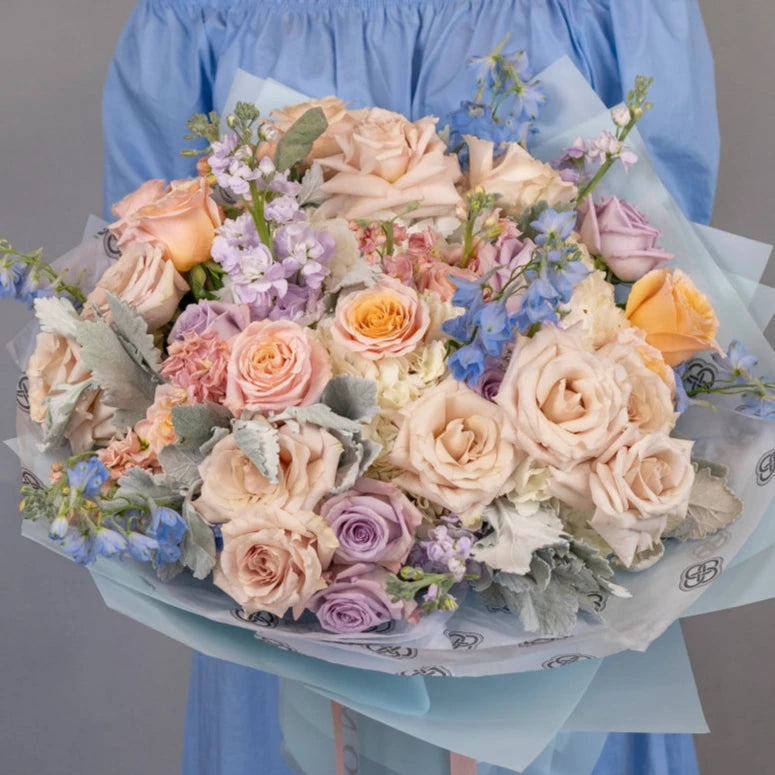 Mixed flower bouquet with peach roses, purple lavender blooms, blue delphinium and dusty miller in white paper