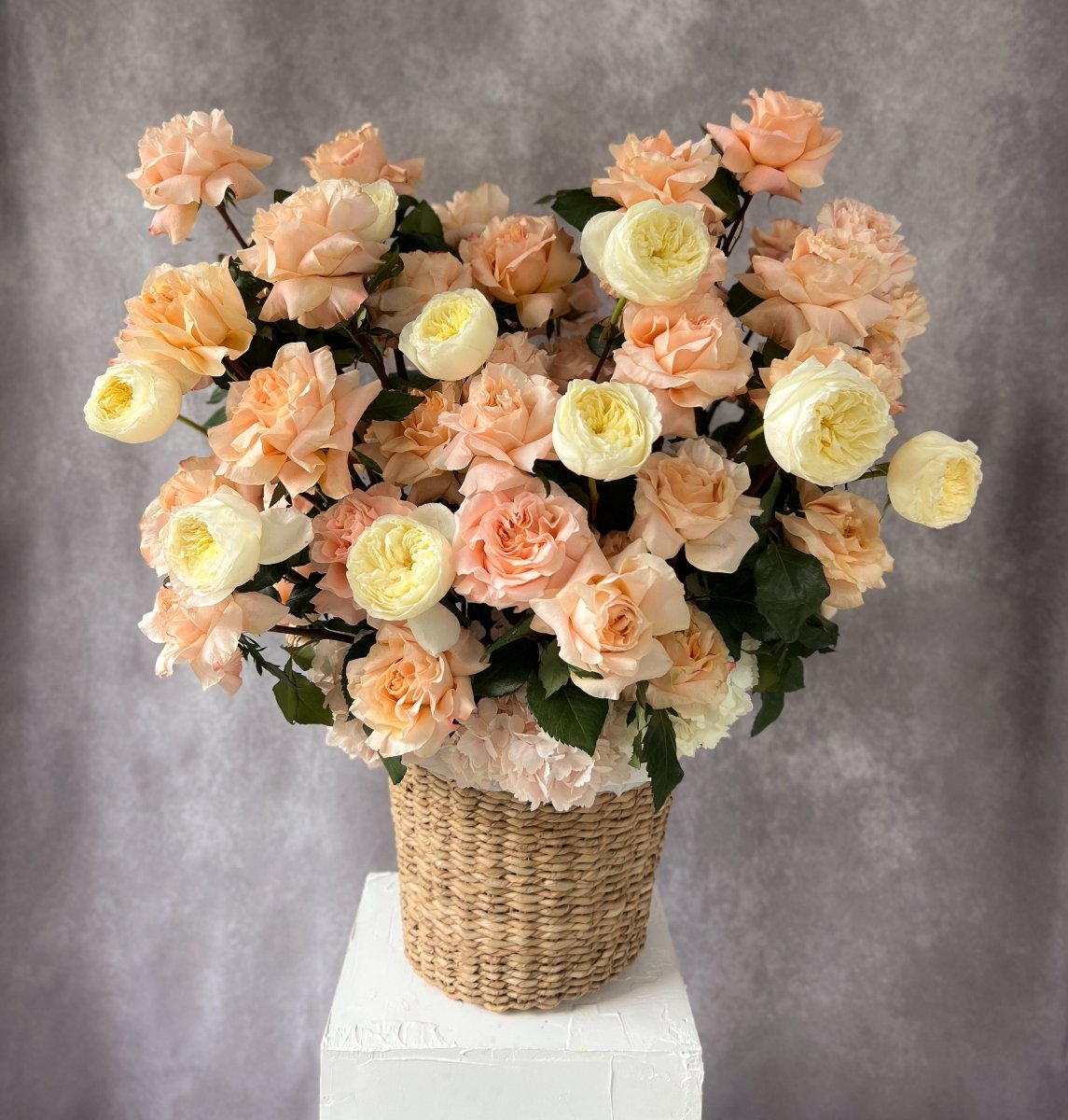 Peach roses and cream ranunculus flower arrangement in woven seagrass basket on white marble surface