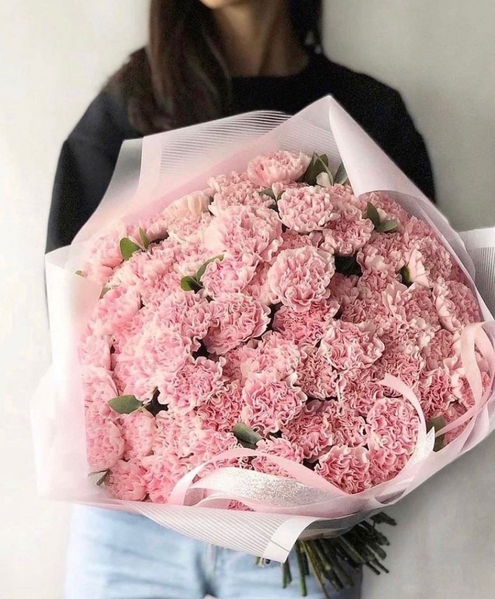 Large bouquet of pink carnations wrapped in translucent pink paper with ribbon, held by woman in black sweater