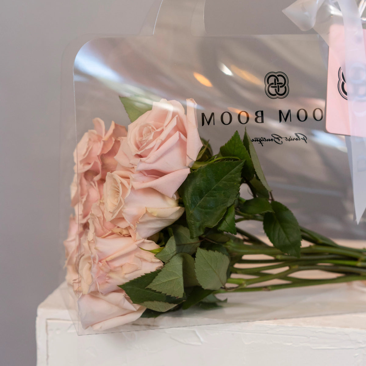 Bouquet of soft pink roses with green leaves and stems next to white Bloom Room boutique shopping bag
