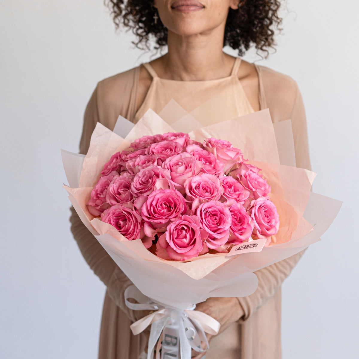 Large pink rose bouquet wrapped in soft peach paper with ribbon, held by woman in beige dress