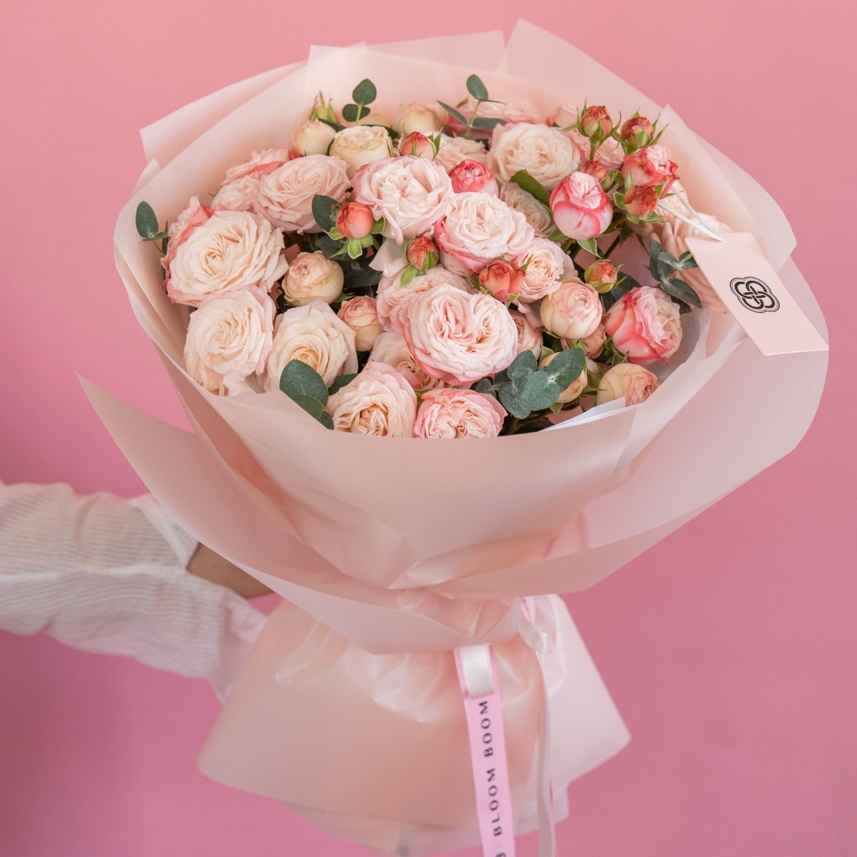 Pink spray rose bouquet with cream and coral blooms wrapped in white paper against pink background