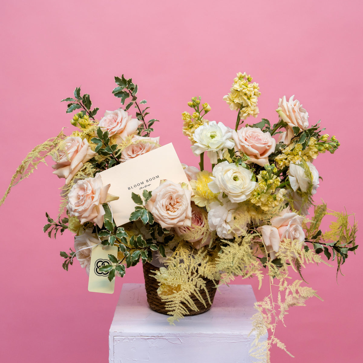 Mixed flower arrangement with pink roses, white ranunculus, and golden ferns in wicker basket on pink background
