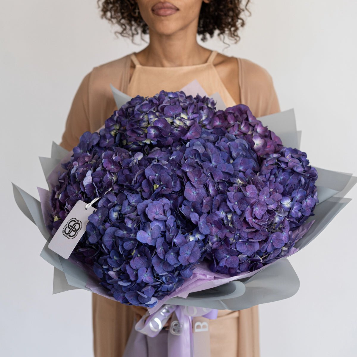 Large purple hydrangea bouquet wrapped in gray paper with white tag held by woman in beige dress
