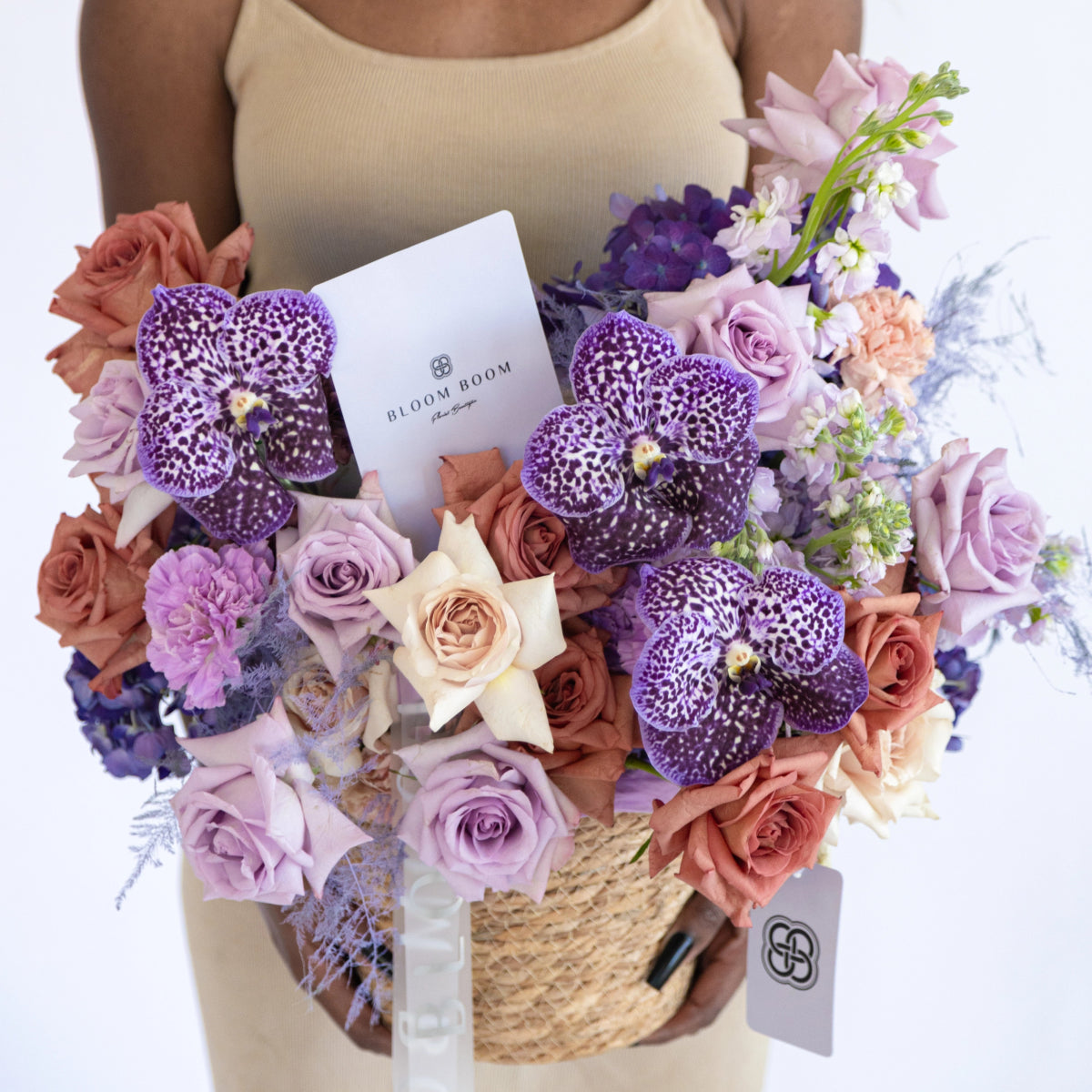 Purple spotted orchid bouquet with lavender roses in woven basket, featuring mixed blooms and Bloom & Boom card