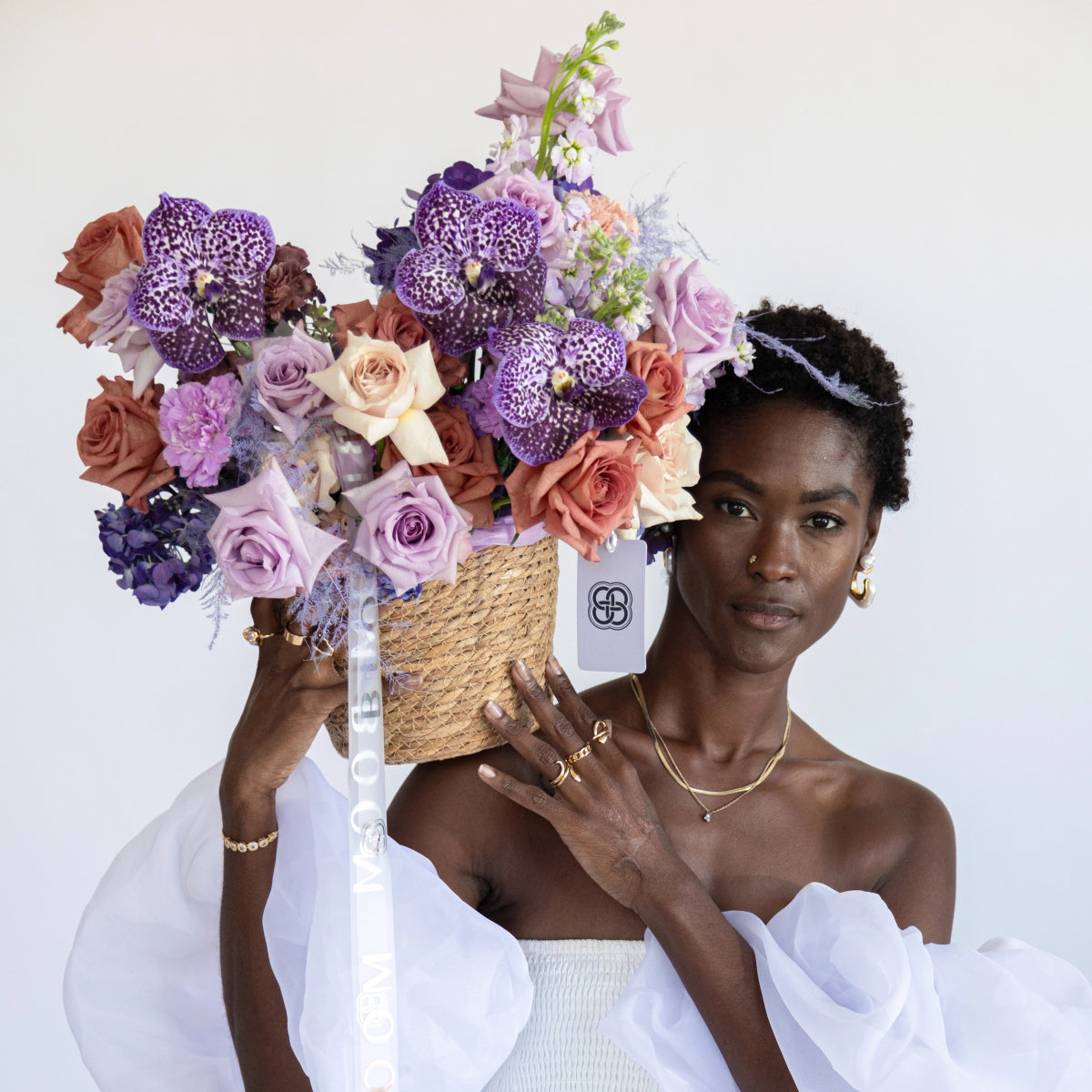Woven basket filled with purple spotted orchids, lavender roses, coral roses and mixed flowers with ribbon