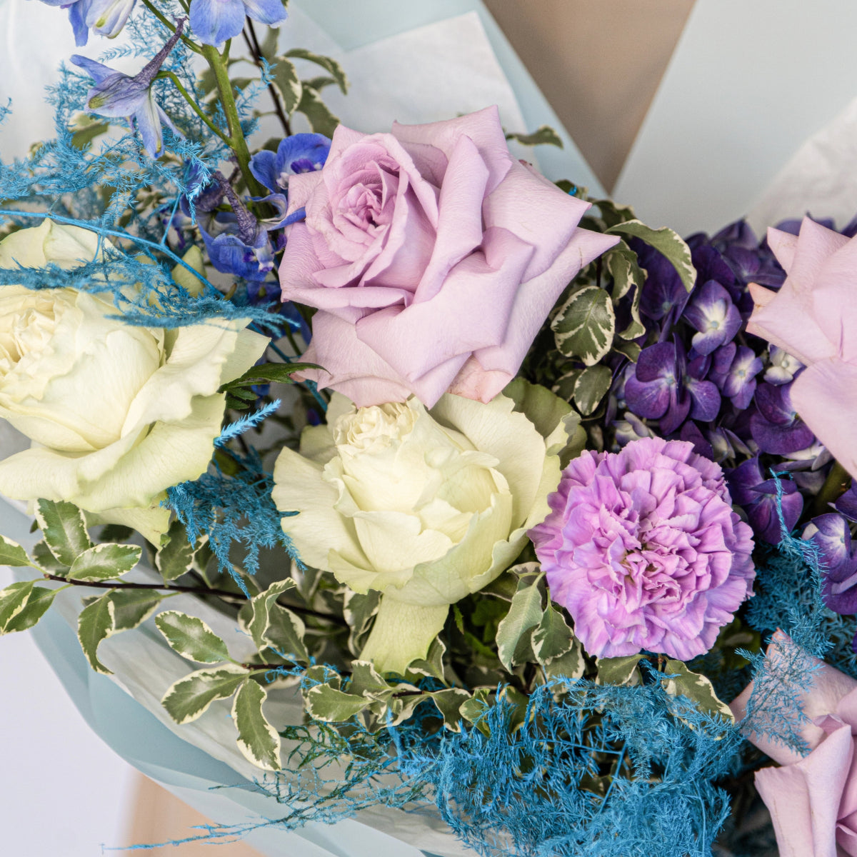 Purple and white rose bouquet with lavender carnation, blue decorative moss, and green foliage in pastel wrapping