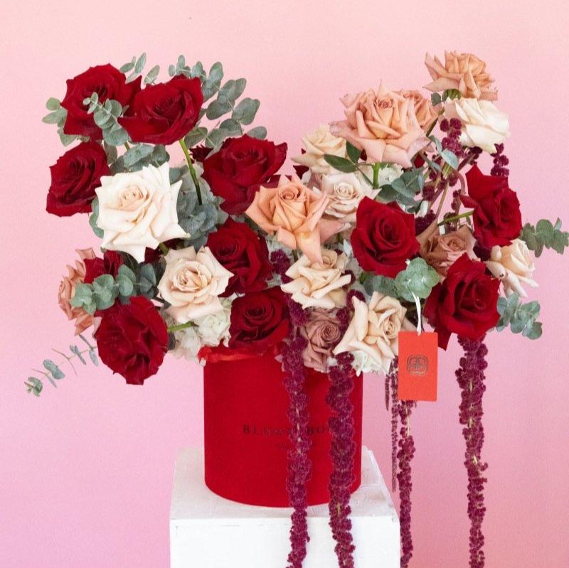 Luxury red hat box filled with deep red and peach roses, eucalyptus leaves, and trailing burgundy amaranthus