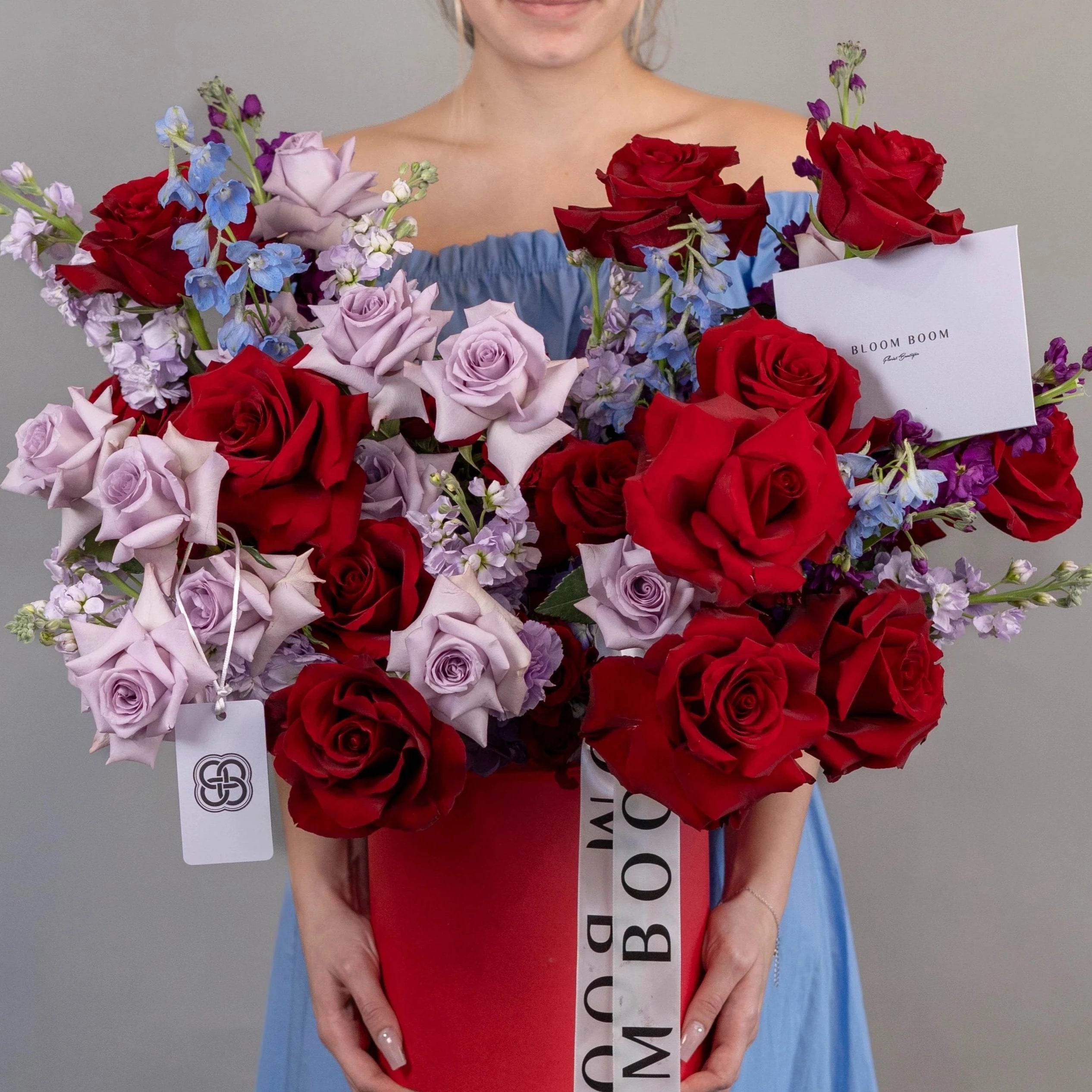 Luxury red box arrangement with red and lavender roses, blue delphinium, and purple stock flowers by Bloom Boom