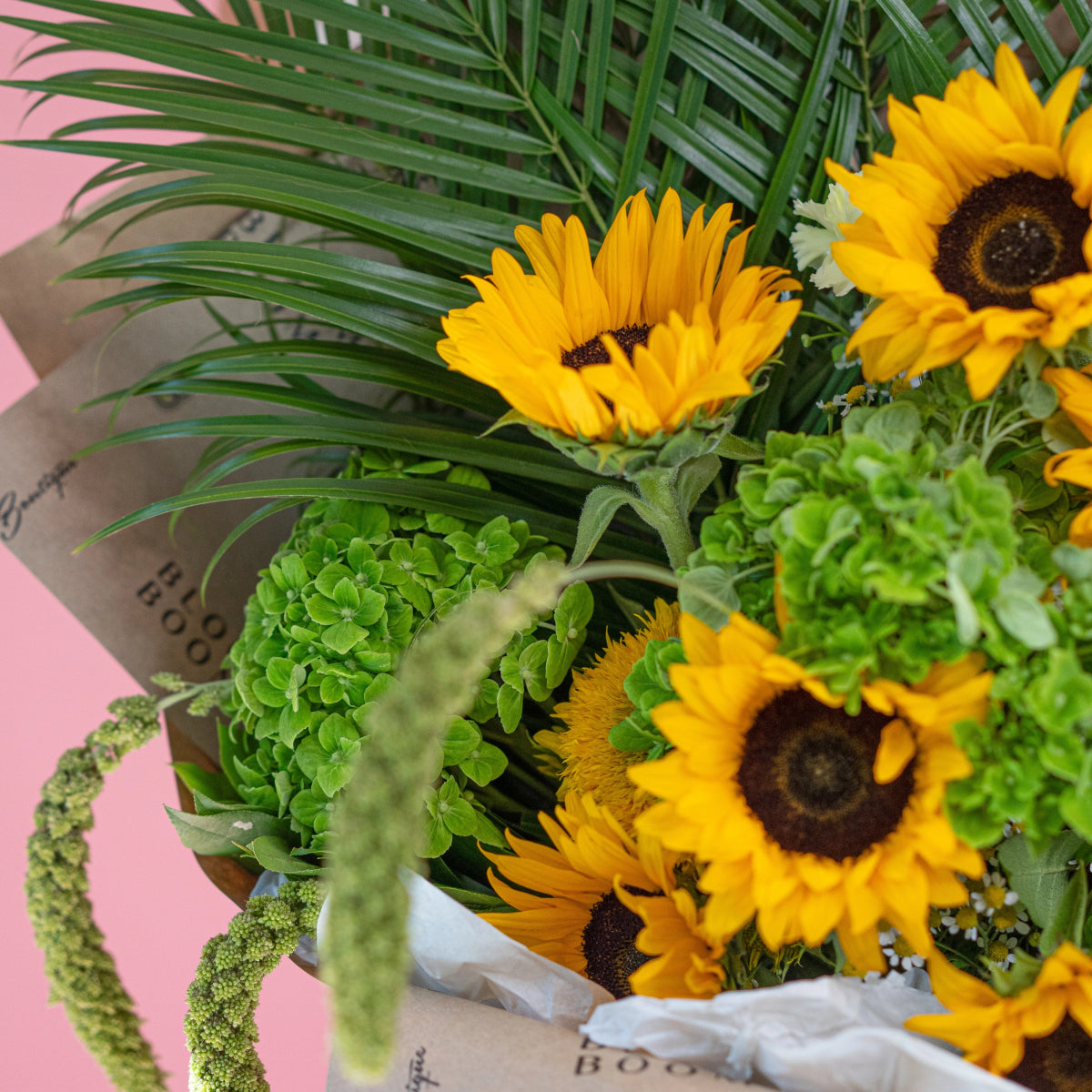 Bright yellow sunflowers with green chrysanthemums and palm fronds in kraft paper wrap with white ribbon