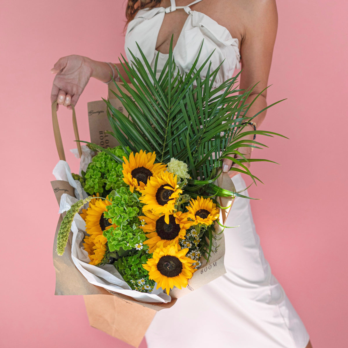 Bright sunflower bouquet with green hydrangeas and palm fronds in brown paper wrapping held against pink background