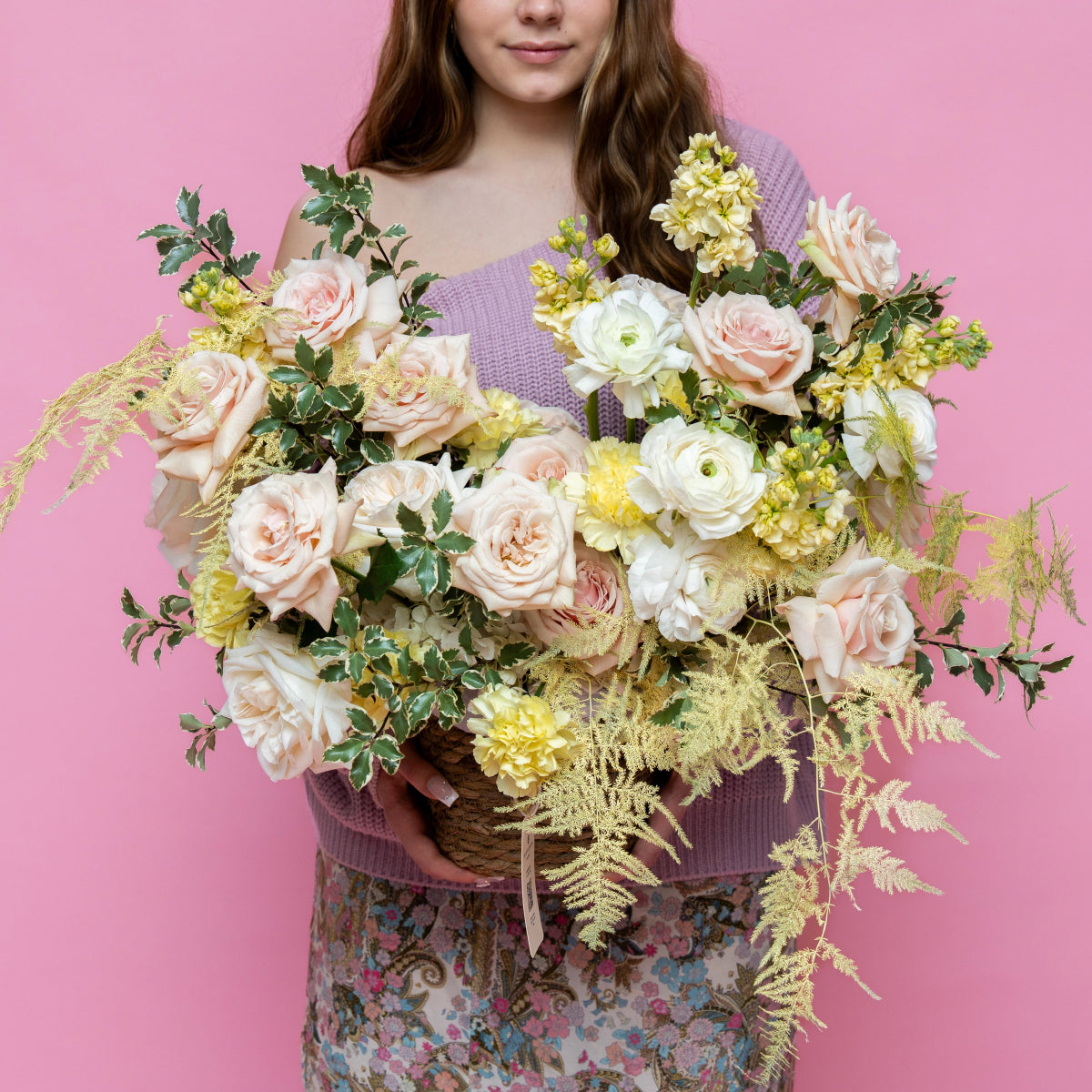 Large wicker basket bouquet with blush pink roses, white ranunculus, and golden ferns against pink background