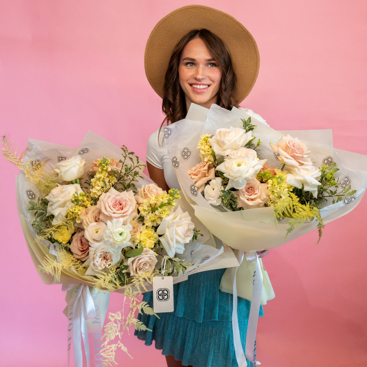 Smiling woman in tan straw hat holding two flower bouquets with peach roses, white ranunculus, yellow blooms
