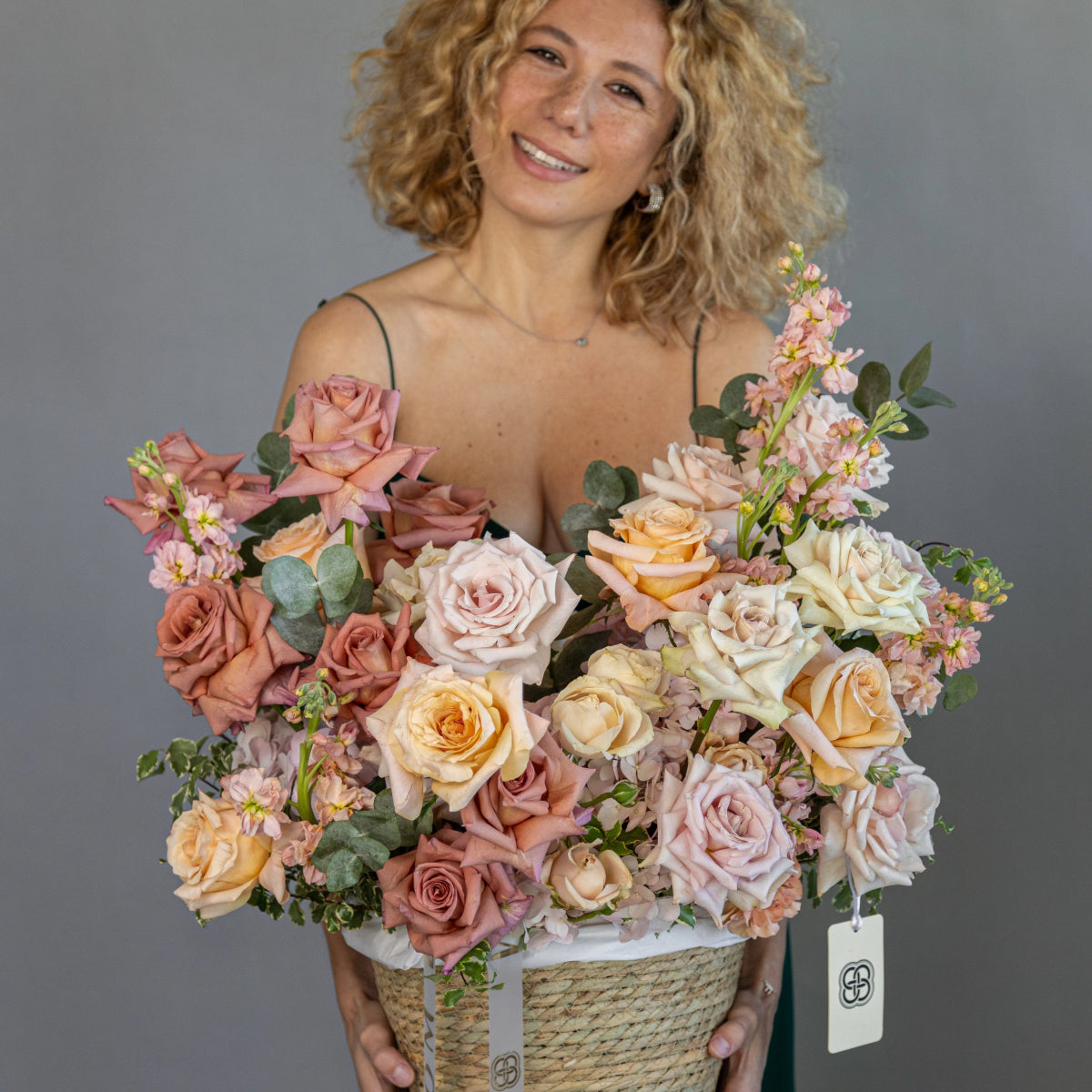 Vanilla Sky flower basket with blush pink and cream roses, eucalyptus in woven rope basket held by smiling woman