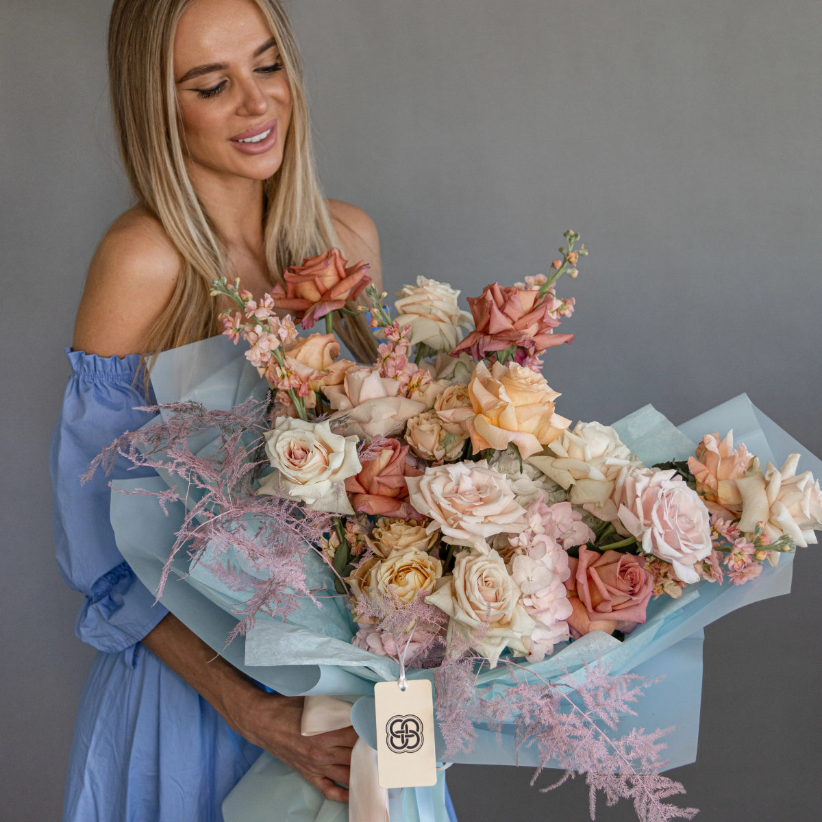 Luxurious mixed rose bouquet in peach, pink, and cream tones wrapped in soft blue paper with decorative tag