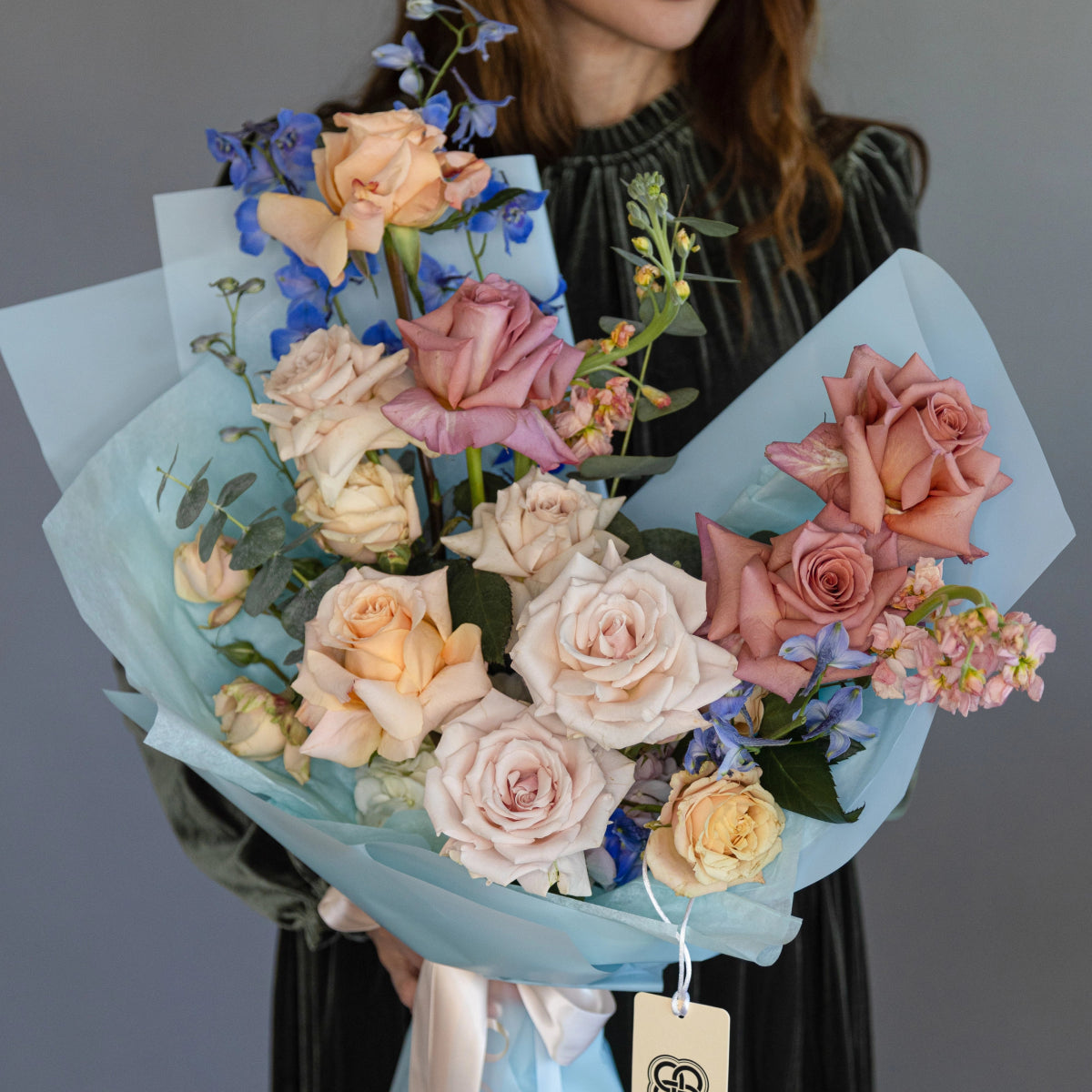 Mixed flower bouquet with peach and blush roses, blue delphiniums, wrapped in light blue paper