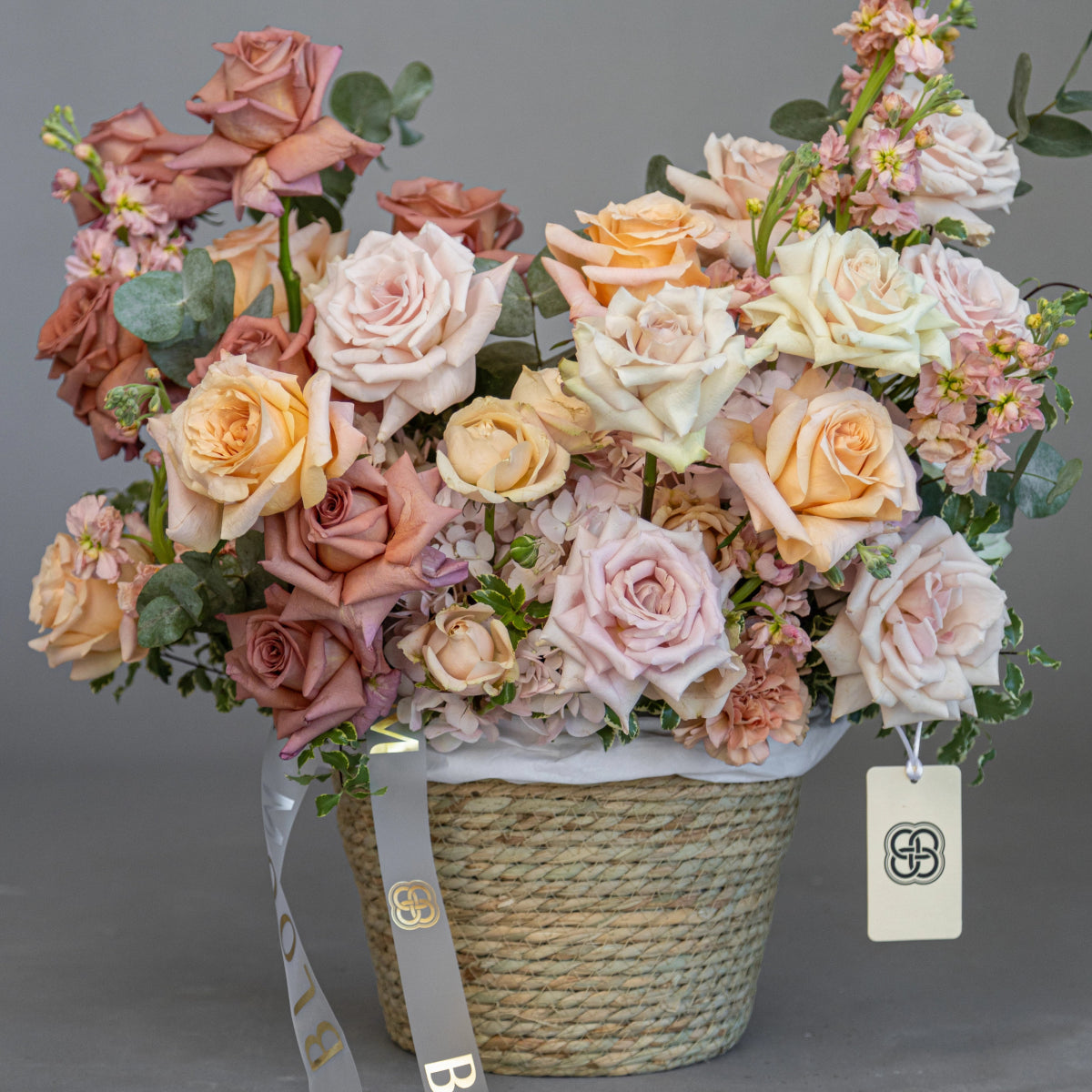 Pastel rose bouquet in woven basket with peach, blush pink and cream roses, eucalyptus and ribbon bow