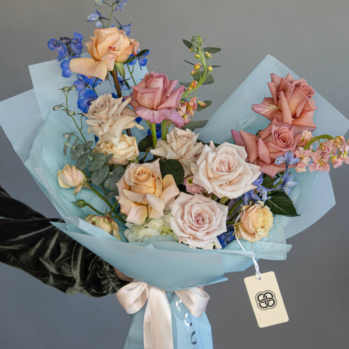 Mixed rose bouquet in blue paper wrap with peach, pink and cream roses, blue delphiniums and gift tag