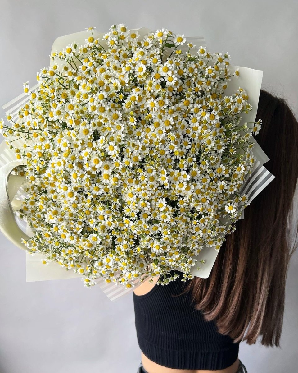 Large white chamomile daisy bouquet with yellow centers wrapped in cream paper, held by woman in black top