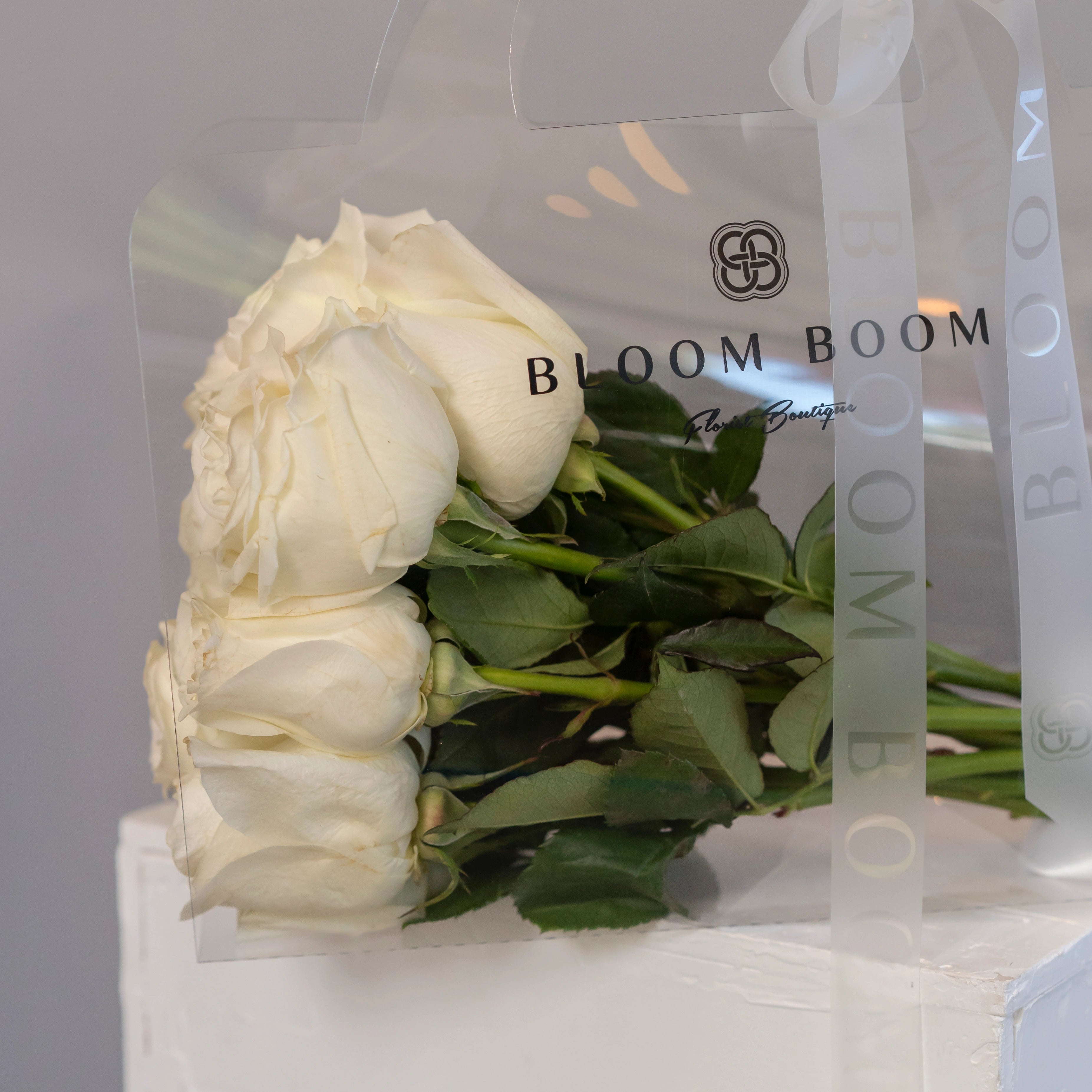 White roses bouquet in clear Bloom Boom branded gift bag with green stems and leaves visible