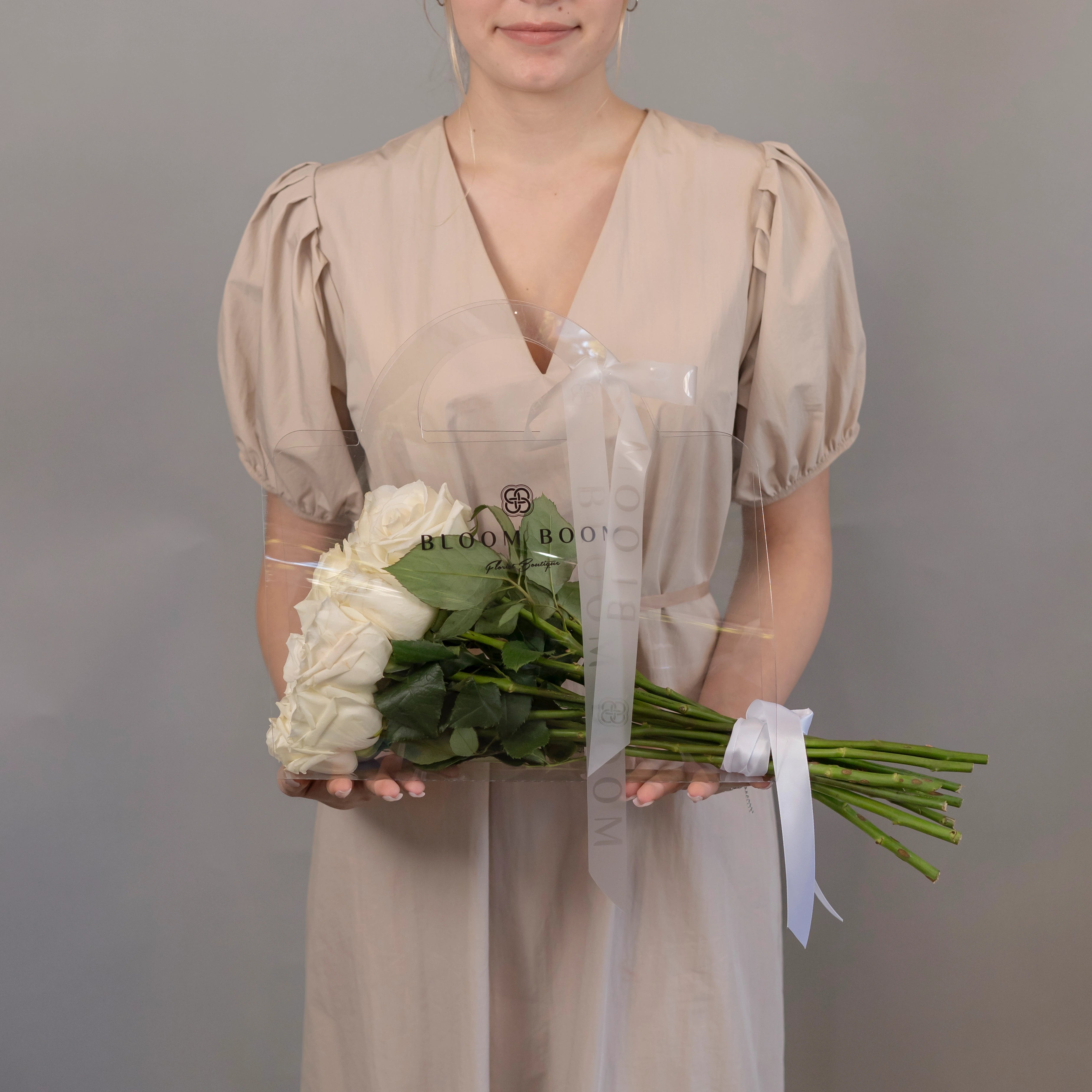 White roses bouquet in clear Bloom Boom branded protective sleeve held by woman in beige dress
